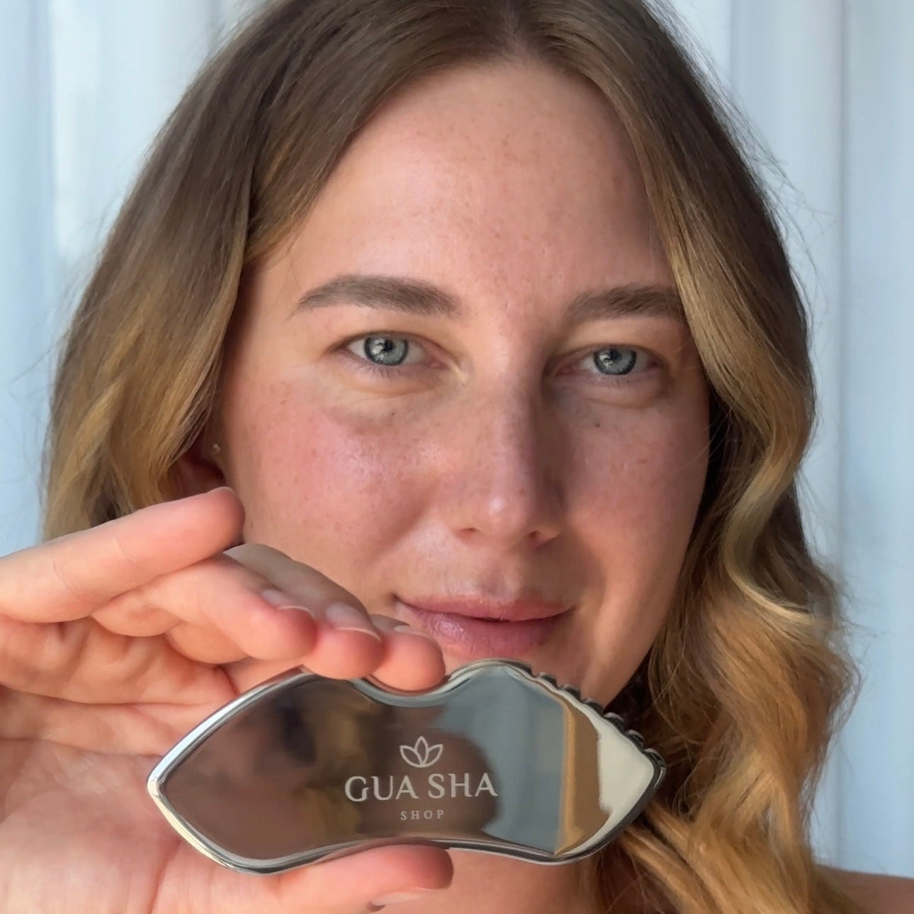 Woman holding a stainless steel Gua Sha tool against her face with a white background